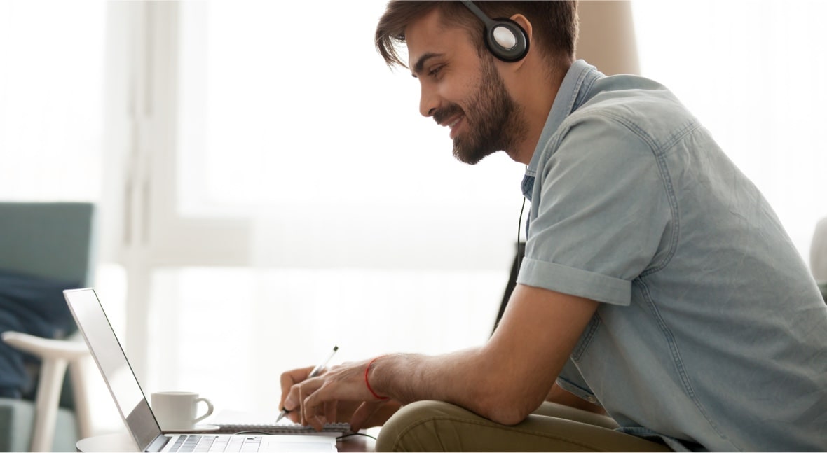 man wearing headphones while taking notes and looking at laptop